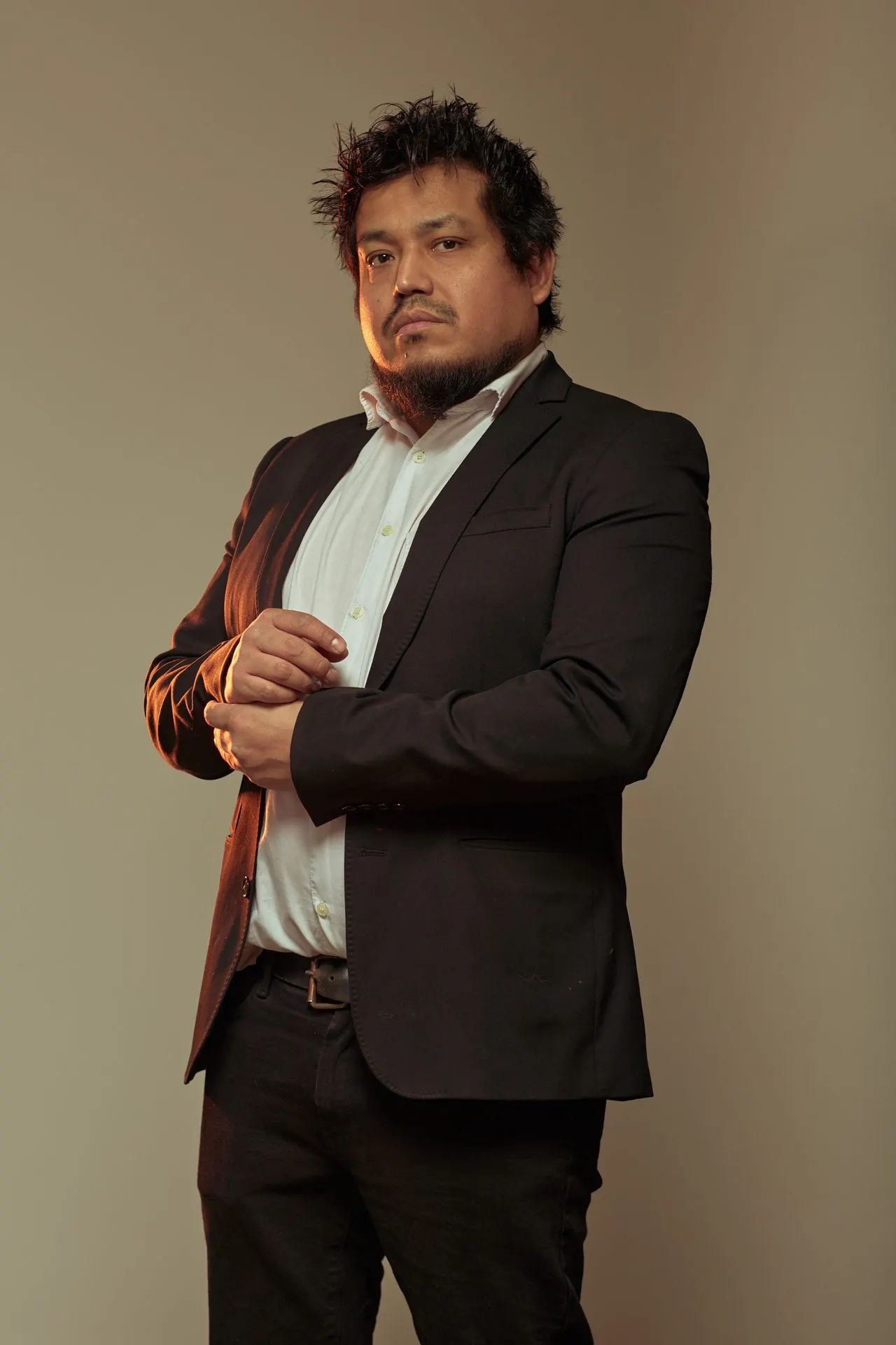 Portrait d'un homme barbu en blazer foncé ajustant sa manchette, fond beige, Bruxelles
