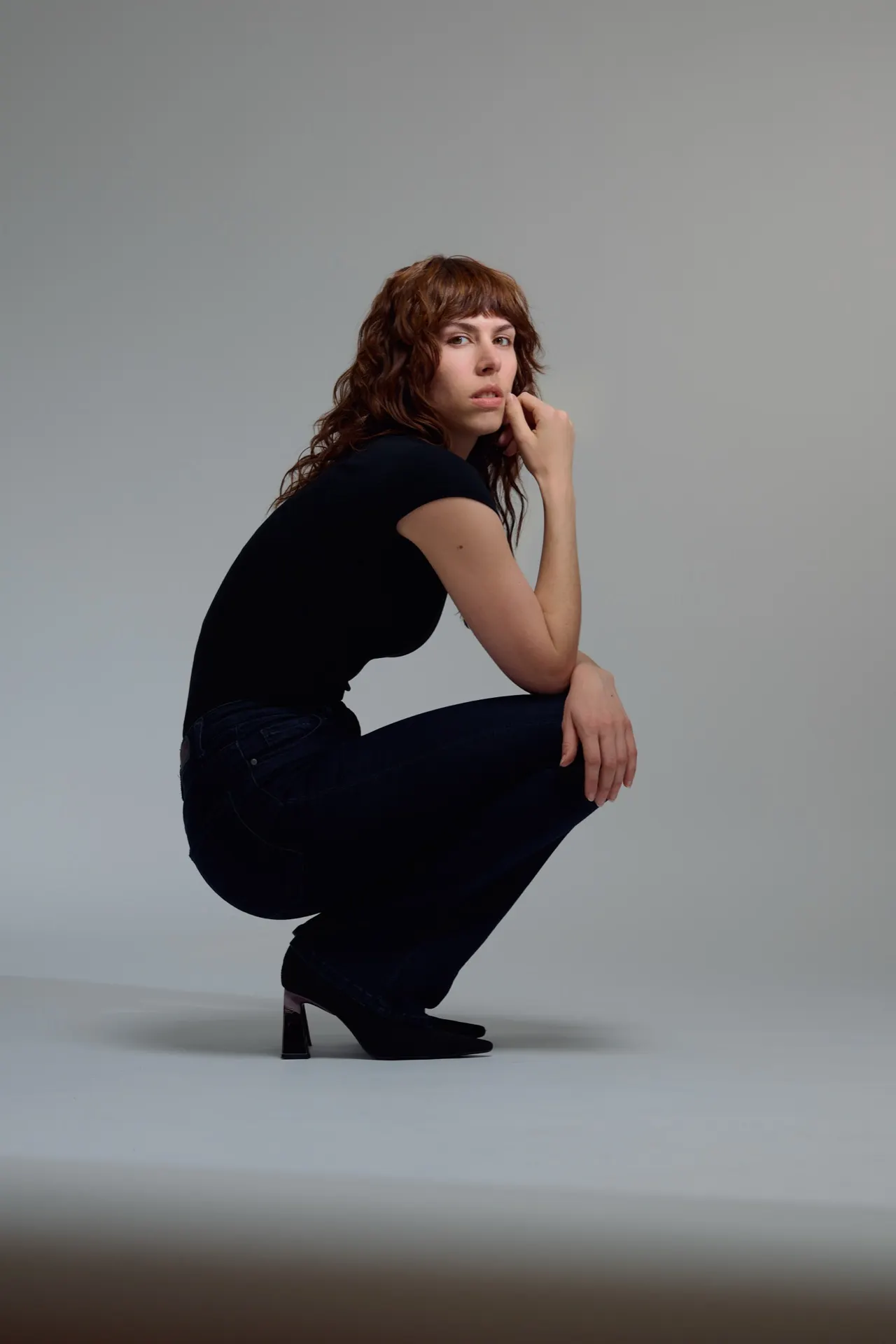 Femme accroupie en t-shirt noir et jean à talons, studio gris, Bruxelles