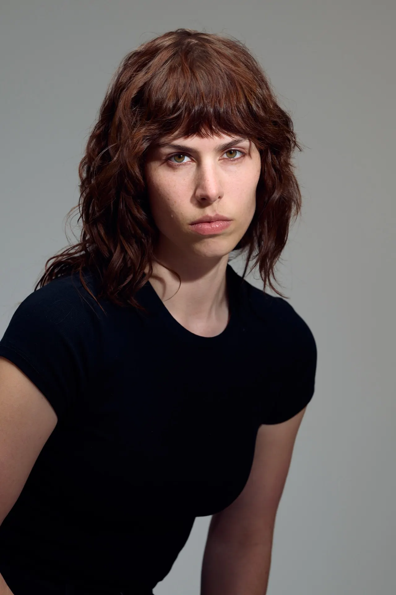 Femme aux cheveux auburn frangés en t-shirt noir, regard intense, studio gris, Bruxelles