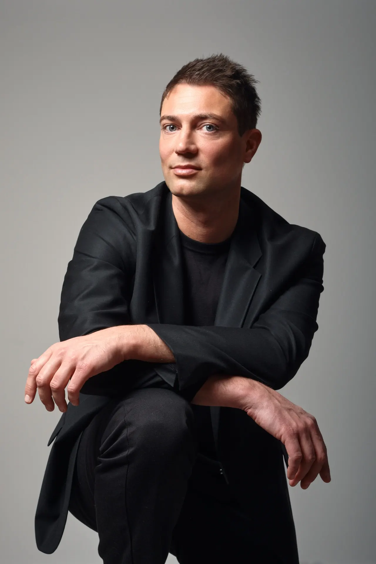Homme en costume noir accroupi, bras croisés, portrait de studio gris, Bruxelles