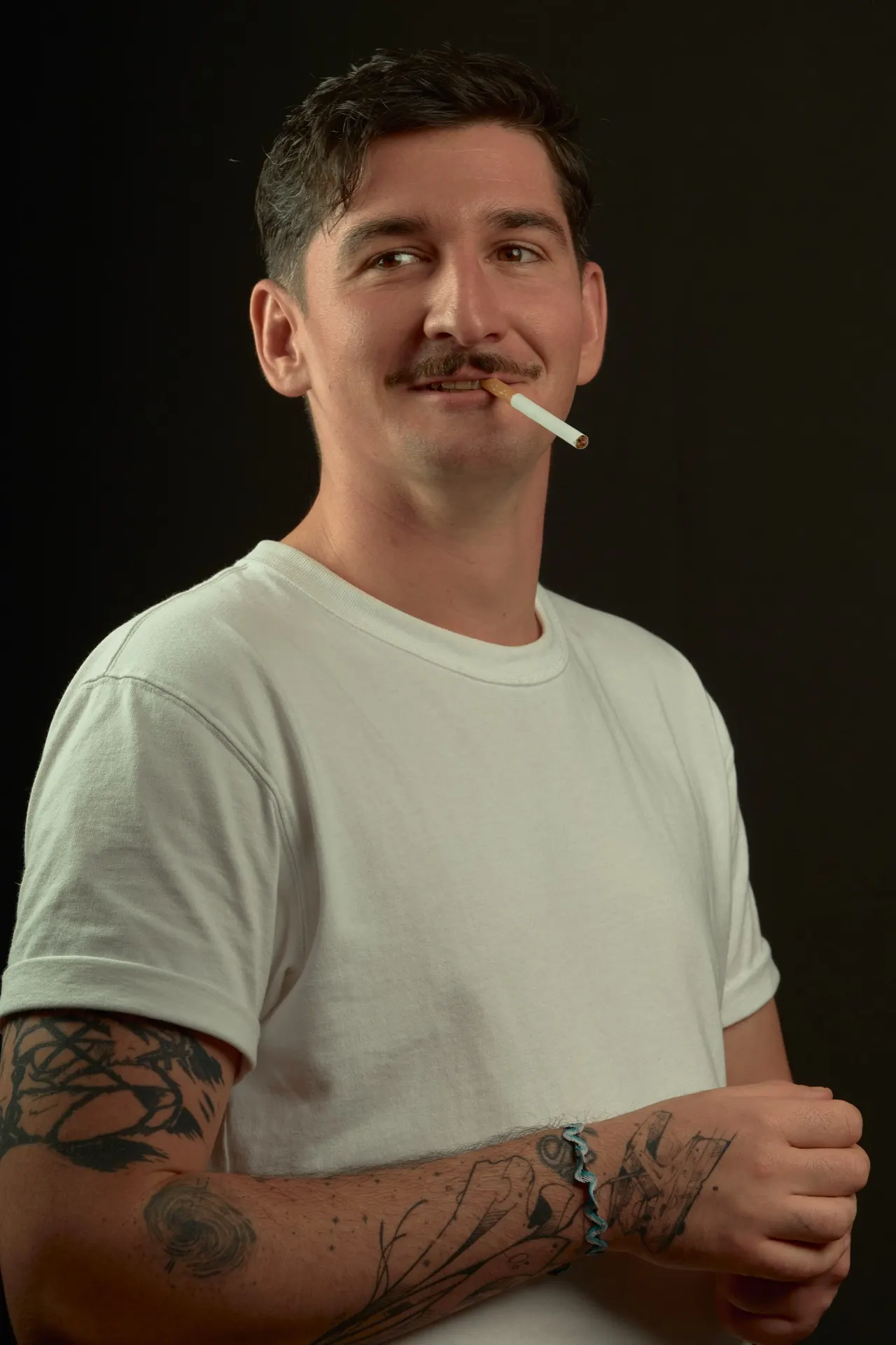 Homme moustachu en t-shirt blanc, cigarette aux lèvres, fond sombre, Bruxelles